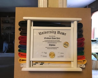Op de afbeelding: Een witte houten display case met een diploma en vechtsportriemen in verschillende kleuren. Het diploma zit in een witte lijst en de riemen zijn aan weerszijden gestapeld. De riemen zijn onder meer zwart, bruin, rood, blauw, groen, oranje, geel en wit.