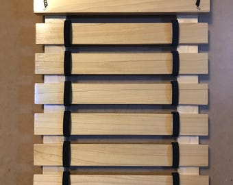 Op de afbeelding: Een houten gordelrek met de naam "ANYA" gegraveerd op de bovenkant. Het rek heeft zes vakken voor gordels en twee zwarte silhouetten van karatefiguren aan weerszijden van de naam.