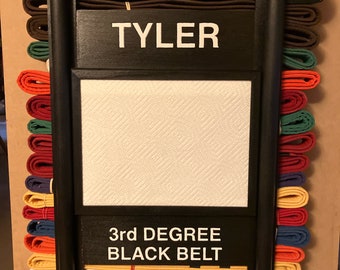 Op de afbeelding: Een zwarte houten gordelrek met een witte achtergrond en de tekst "TYLER 3rd DEGREE BLACK BELT". Het rek is gevuld met een verscheidenheid aan gekleurde gordels.