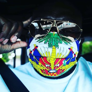 May include: A person wearing a face mask with the flag of Sint Maarten. The mask is white with a yellow, blue, and red design. The person is also wearing gold-rimmed sunglasses.