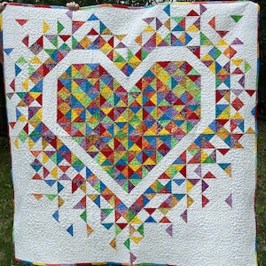 May include: A white quilt with a large heart-shaped design made of colorful patchwork triangles. The heart is surrounded by a border of smaller triangles.