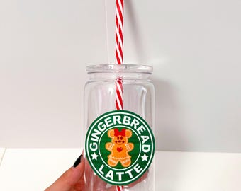 May include: Clear glass tumbler with a green and white sticker that says "Gingerbread Latte" with a cartoon gingerbread person wearing a red bow. A red and white striped straw is in the tumbler.