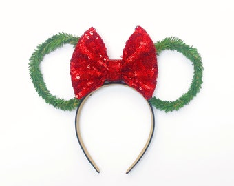 May include: A festive headband featuring a large, sequined red bow and green wreath-shaped ears. The headband is black and the bow is centered above the band. The ears are made of green faux evergreen branches.