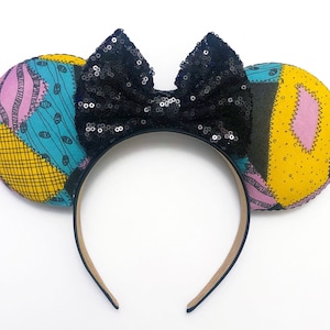 May include: Black and white patchwork fabric mouse ears with a black sequin bow. The headband is beige.