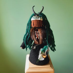 Peut inclure: Poupée artisanale avec des dreadlocks verts, une robe noire et un col en fourrure. La poupée présente une coiffe avec des cornes, un masque à franges et des perles décoratives. Une parure de plumes et de perles est suspendue sur le côté.