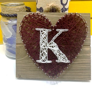 May include: A wooden sign with a heart-shaped design made with red and white string wrapped around nails. The letter "K" is spelled out in white string within the heart.