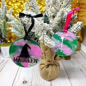 May include: Two round Christmas ornaments with green, pink, and silver swirls. One features a black witch hat and the name "MAUREEN". The other has a white design and the name "Sally". Both have ribbons for hanging.