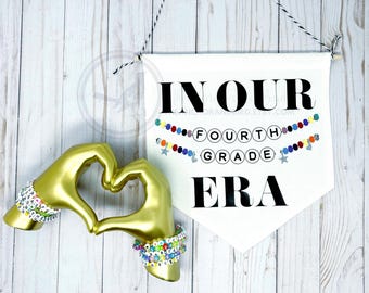 Pode incluir: Faixa branca com texto preto que diz "In Our Fourth Grade Era". A faixa é decorada com contas coloridas e estrelas prateadas. Duas mãos douradas formam um coração na frente da faixa.