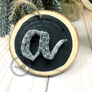 May include: A silver wire wrapped around nails to form the letter "a" on a black painted wooden round ornament. The ornament is attached to a twine string.