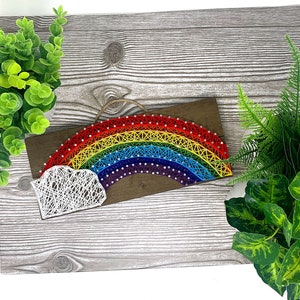 May include: A rainbow string art piece with a white cloud on a wooden background. The rainbow is made of colorful yarn and the cloud is made of white yarn.