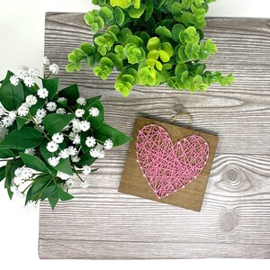 May include: A pink heart-shaped string art piece on a wooden background. The heart is made of pink thread wrapped around nails.