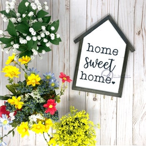 May include: A wooden sign with a white background and black text that reads "home sweet home". The sign has a distressed look and is hanging on a white wooden wall. There are flowers in the background.
