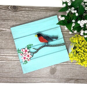 以下が含まれることがあります： ピンクの花が咲いた枝に、赤と黒の鳥がとまっている青い木製の看板。看板は木の背景の上に置かれています
