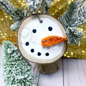 May include: A wooden snowman ornament with a white painted face, black eyes and mouth, and an orange yarn nose. The ornament is attached to a burlap ribbon.