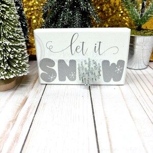 May include: White wooden block sign with the text "Let it Snow" in gray lettering. A silver glitter snowflake is attached to the sign.