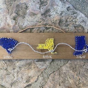 May include: A wooden sign with three string art state outlines. The states are South Carolina, New York, and Massachusetts. The dates 5/14/13, 4/19/16, and 7/16/17 are written below each state.