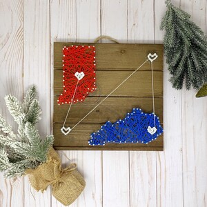 Puede incluir: Un letrero de madera con arte de hilo que representa los estados de Indiana y Kentucky. Los estados están conectados por un hilo blanco con un corazón al final. El letrero está sobre un fondo de madera blanco.