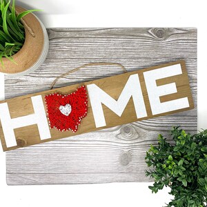 May include: A wooden sign with the word "HOME" in white letters. The letter "O" is replaced with a red string art design of the state of Ohio with a white heart in the center.