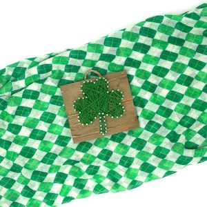 Könnte beinhalten: Ein grünes Kleeblatt aus String Art auf einem Holzgrund. Das Kleeblatt liegt auf einem grün-weiß karierten Stoff.