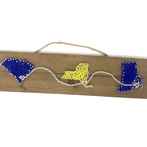 May include: A wooden sign with string art depicting the outlines of the states of South Carolina, New York, and Connecticut. The string art is done in blue, yellow, and white thread.