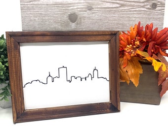 May include: A framed black and white line art print of a city skyline. The frame is dark brown wood and the print is on a white background.