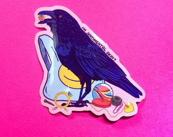 May include: A black crow with a blue tint holding a small pink and gold object in its beak. The crow is standing on a pink background with a blue and yellow bag, a gold ring, a screw, a paperclip, a button, a black nut, a marble, and a yellow rubber ducky. The text "I'M SENTIMENTAL OKAY?" is written on the bag.