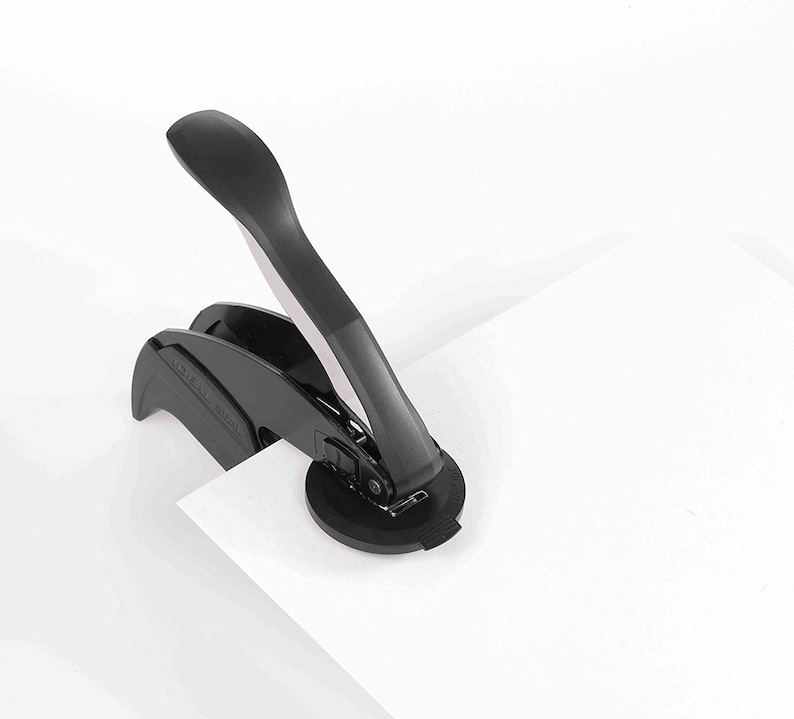 May include: A black desk embosser with a long handle and a circular base, placed on a white sheet of paper. The embosser is designed to create raised impressions on paper. The handle is angled upwards, suggesting it's ready for use.