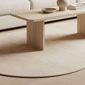 Puede incluir: Escena de salón de tonos neutros con una mesa de centro de madera clara, un sofá color crema y una alfombra redonda. Un jarrón con vegetación y un cuenco pequeño están sobre la mesa. Un cojín texturizado descansa en el sofá.