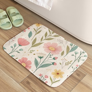 Könnte beinhalten: Eine florale Badematte mit cremefarbenem Hintergrund und einem Muster aus rosa, gelben und grünen Blumen und Blättern. Die Matte liegt auf einem Holzboden neben einer weißen Badewanne und einem Paar hellgrüner Sandalen.
