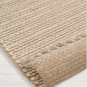 Peut inclure: Gros plan d'un tapis tiss&eacute; beige et blanc avec une bordure &agrave; franges. Le tapis est roul&eacute;, montrant la texture du tissage.