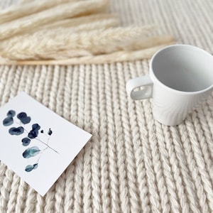 A close-up view of a hand-knitted chunky wool area rug, showcasing its thick, ivory fibers. The soft, felted wool has a modern, flatweave design that adds texture and warmth to the space.