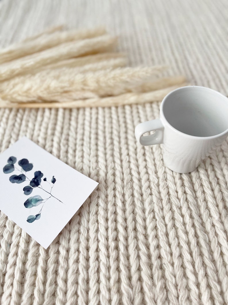 A close-up view of a hand-knitted chunky wool area rug, showcasing its thick, ivory fibers. The soft, felted wool has a modern, flatweave design that adds texture and warmth to the space.