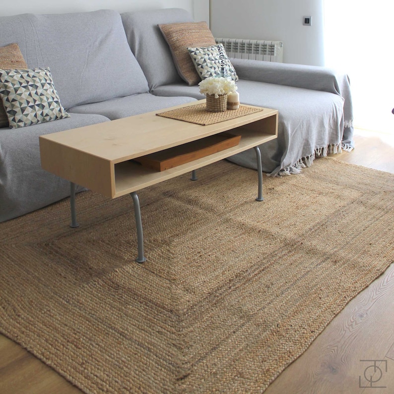 A light brown jute rug with a woven pattern. A light wood coffee table with a shelf sits on top of the rug. The coffee table has silver legs.