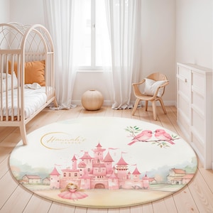 Puede incluir: Una alfombra redonda de color crema con una ilustración de un castillo rosa y el texto "Hannah's Castle" en dorado. La alfombra también incluye una bailarina y dos pájaros rosas en una rama, ideal para una habitación infantil.