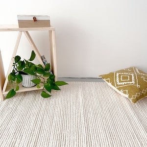 A spacious living room featuring a hand-woven wool flatweave rug. The rug is ivory, with a chunky knit pattern that creates a pebble-like texture. Its thick, soft wool enhances the cozy atmosphere, complementing the room's modern decor.