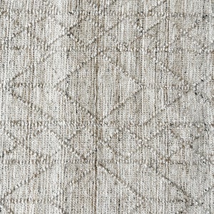 May include: Close-up of a white and beige woven rug with a diamond pattern. The rug has a textured surface and a subtle sheen.
