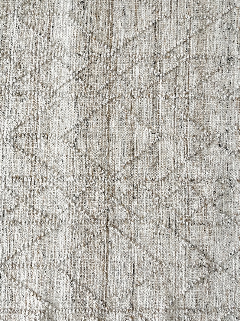May include: Close-up of a white and beige woven rug with a diamond pattern. The rug has a textured surface and a subtle sheen.