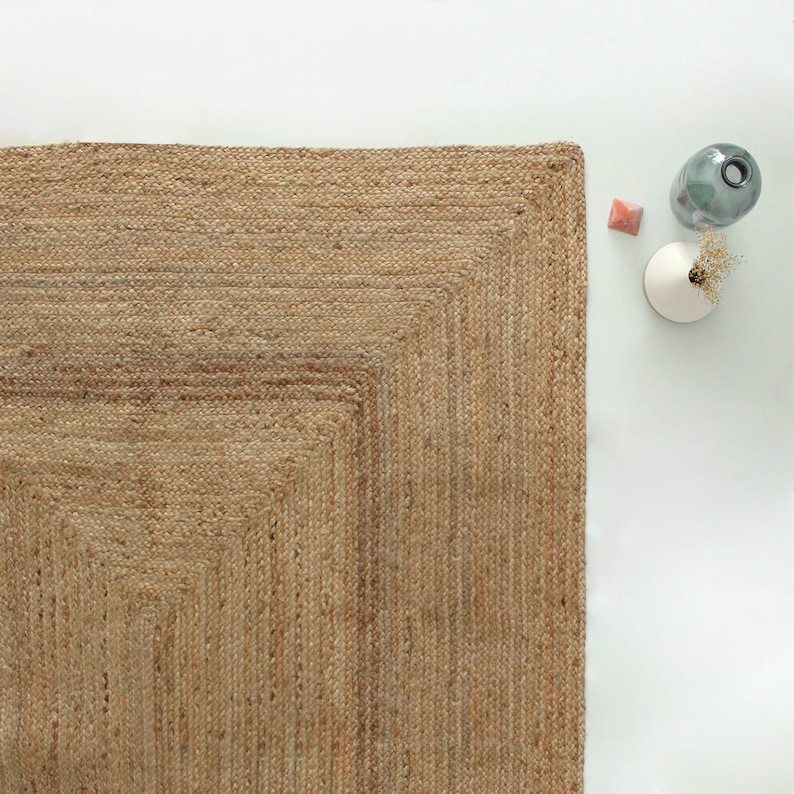 A natural fiber rug with a woven texture in a light brown color. The rug has a square shape with a border that is slightly darker than the center of the rug.
