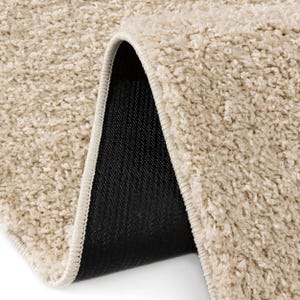 May include: Close-up of a beige, shaggy rug with a rolled-up corner. The rug has a soft, textured surface and a cream-colored border. The underside is black, providing a contrast to the light-colored top.
