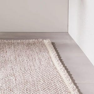May include: A rectangular, neutral-toned rug with a textured, woven appearance. The rug features a light beige and gray color palette, with a white border along one edge. The rug is placed on a gray surface.