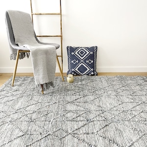 May include: A gray and white woven rug with a diamond pattern. The rug is in a living room setting with a chair and a pillow.