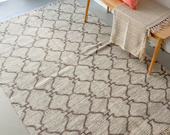 May include: A cream-colored rug with a brown geometric pattern and fringe edges. The rug is laid out on a gray floor with a wooden bench and pillows in the background.