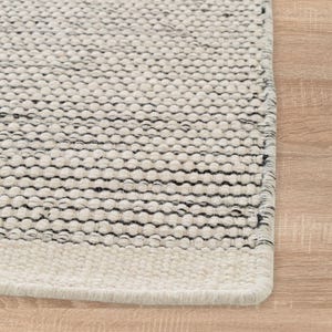 May include: Close-up of a white and black woven rug with a textured surface. The rug has a fringe border.