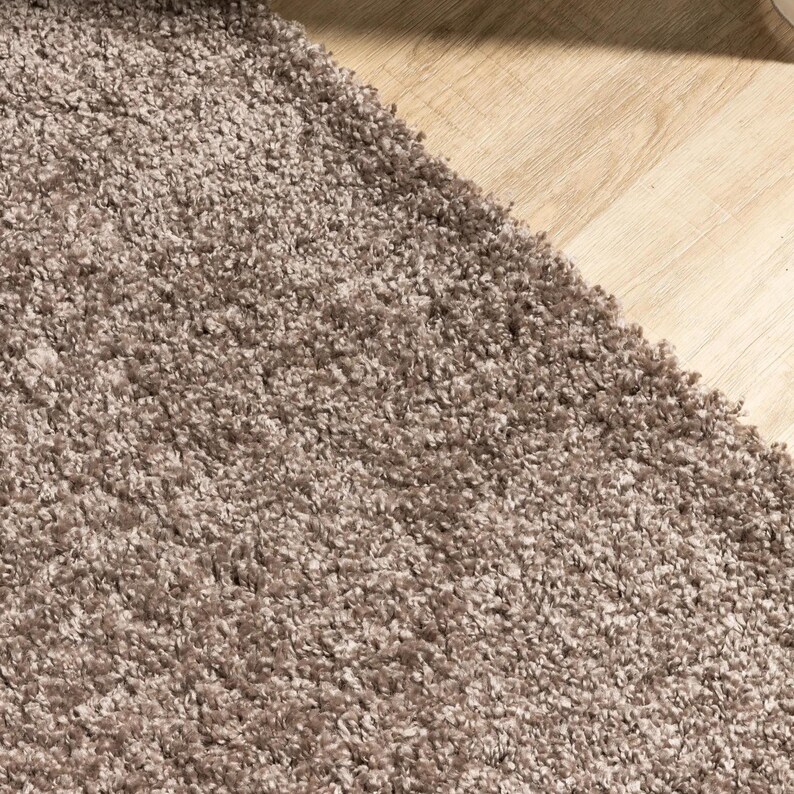 May include: Close-up of a plush, textured rug in a neutral taupe color. The rug has a dense, shaggy pile, creating a soft and inviting appearance. The rug's edge meets a light-colored wooden floor.