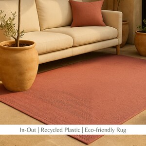 Puede incluir: Una alfombra rectangular de color terracota con una superficie texturizada. La alfombra está colocada frente a un sofá beige con un cojín terracota a juego. Una planta en maceta está a la izquierda. La alfombra es de plástico reciclado.