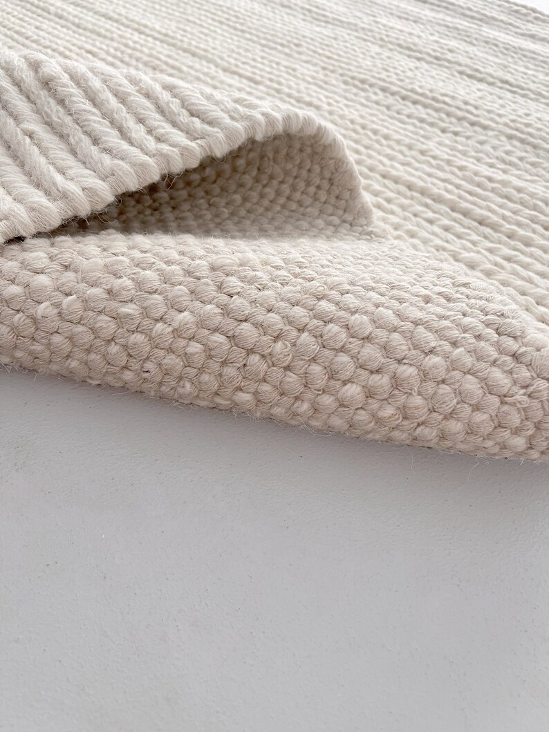 A modern living room showcasing a hand-woven wool flatweave rug. The rug's chunky knit texture and off-white color create a cozy ambiance. Its thick, soft wool fibers add depth to the room's clean, Scandinavian aesthetic.