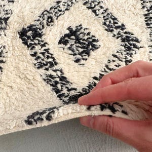 May include: Close-up of a white and black patterned rug with a hand holding the edge. The rug has a fluffy texture and a geometric design.