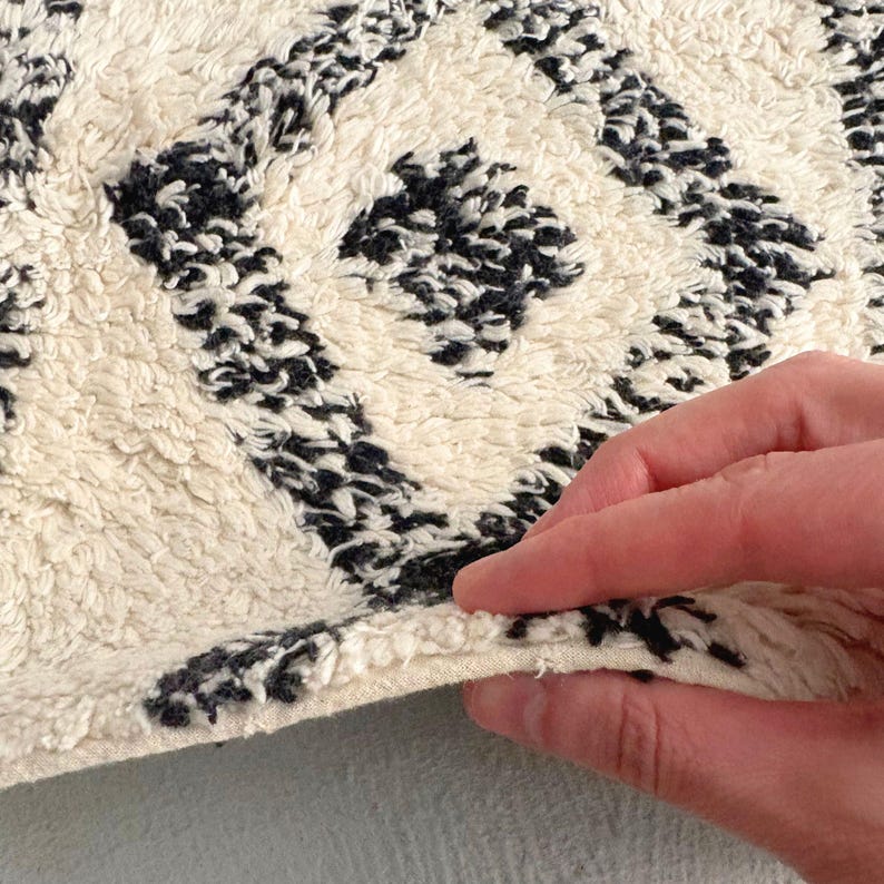 May include: Close-up of a white and black patterned rug with a hand holding the edge. The rug has a fluffy texture and a geometric design.