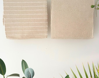 May include: Two beige woven placemats hang against a white wall. One features horizontal stripes, the other a diamond pattern. Green plants frame the scene, adding a natural touch to the neutral tones.