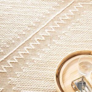 Peut inclure: Gros plan sur un tapis tiss&eacute; avec un motif g&eacute;om&eacute;trique blanc et beige. Un plateau en bois avec un verre et une petite bo&icirc;te sont pos&eacute;s sur le tapis.